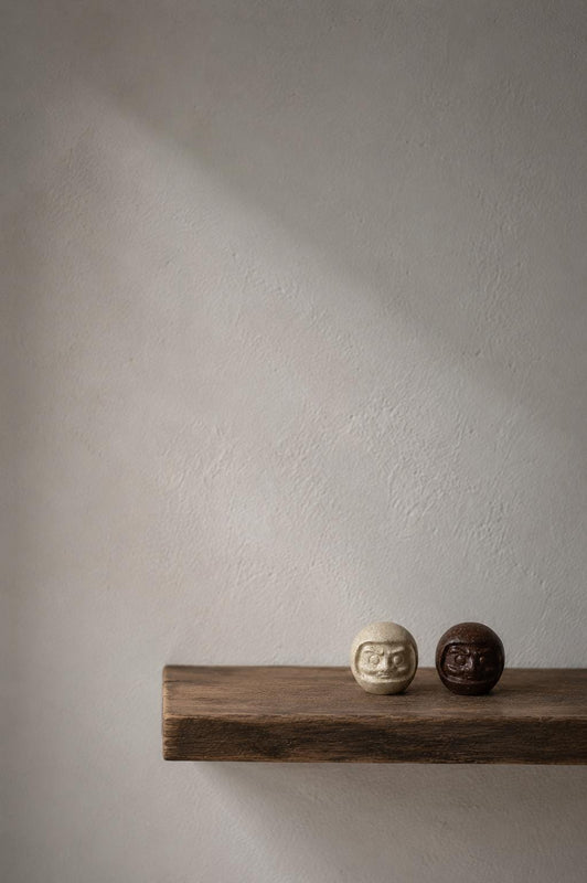 Two small daruma figures on a wooden shelf against a softly lit wall